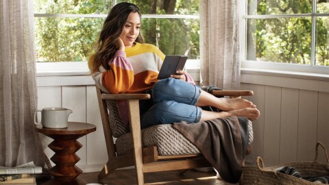 A woman lounges in a chair, with her feet tucked to the side, reading a new Amazon Kindle Paperwhite. 