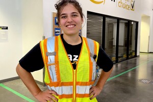 megan coghlan a process assistant at amazon standing in the foreground of an amazon fulfillment center. she is smiling with her hands on her hips and wearing yellow safety vest