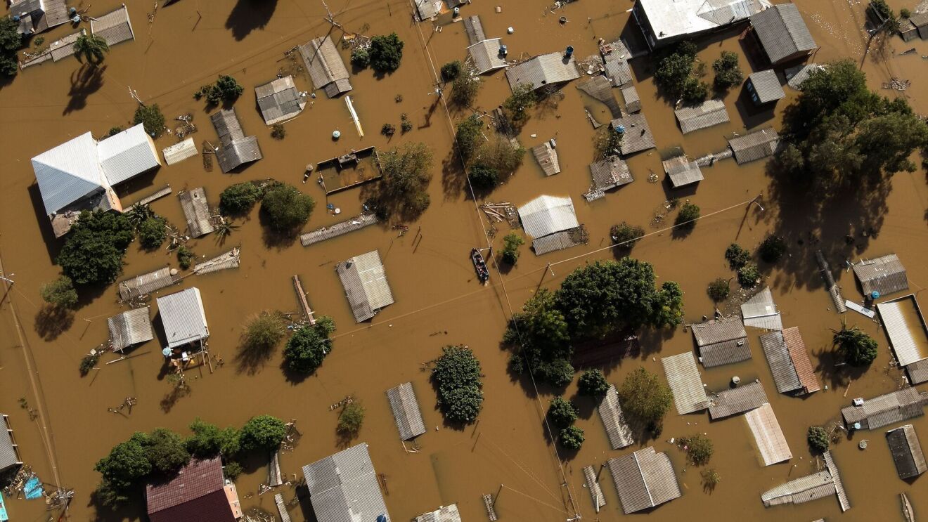 An aerial view of a town in Brazil that is flooded. The roofs of buildings are visible and surrounded by brown water.