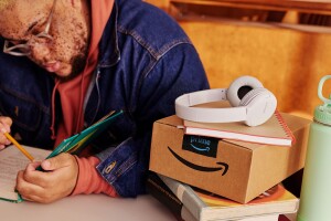 A man in a denim jacket opens a book to study; to his left on the table there's an unopened Amazon box, a mint green Takeya water bottle, and white Sony headphones.