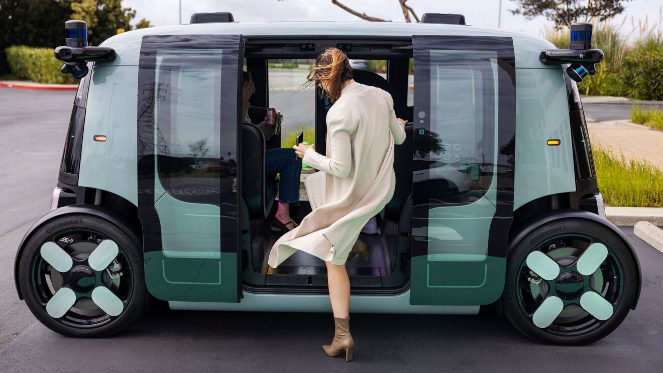 An image showing the Zoox robotaxi opening it's doors on both sides of the vehicle to welcome passengers. There is a passenger entering the vehicle while holding her cellphone and a canned drink. The robotaxi is teal and black and the doors are on a sliding function.