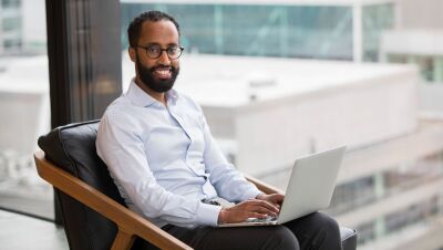 An image of a man seated in front of an office building window with a laptop.