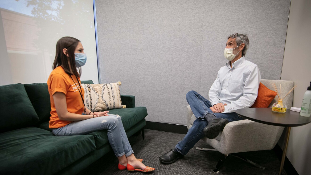 A man and a woman sit in a clinic setting wearing masks while speaking.