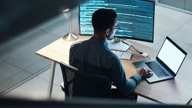 A photo of a developer sitting at a desk. They are working on their laptop device, which is displaying code on a monitor.