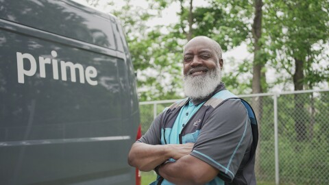 A photo of an Amazon delivery truck driver standing with their arms crossed, in front of a delivery van.