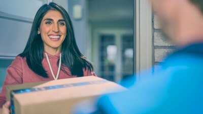 A woman with long dark brown hair smiles as she accept her Amazon package from a delivery driver. 