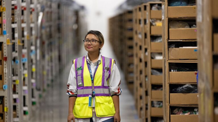 Woman wearing a safety vest on the floor of an Amazon fulfillment center. She's in profile, looking to her righ.