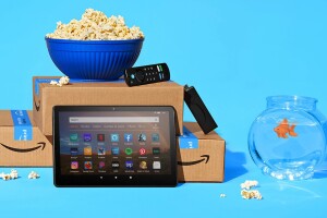 An image of two amazon boxes with a bowl of popcorn on top, a Fire TV stick and remote, and a tablet in front. Next to the boxes is a fishbowl and the background is bright light blue. 