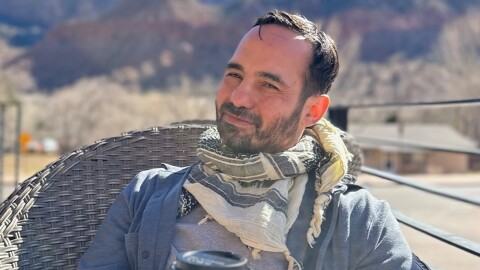 Chief Meteorologist Victor Rodriguez holds a coffee in his hands as he sits in a chair near the base of mountains.