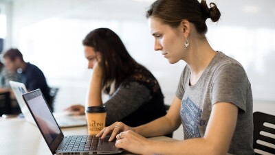 A woman in a t-shirt works on a laptop computer in a workplace setting. Next to her, another woman works on a laptop, and behind them, men sit at a table. 