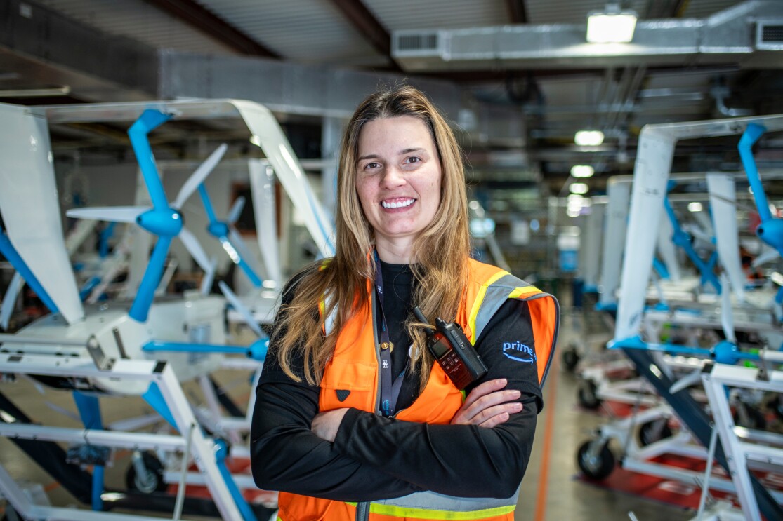 An image of Christina Carter smiling for a portrait at the Amazon drone facility in Texas. There are several rows of drones behind her.