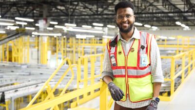 An image of Abdiaziz, an area manager working in an Amazon fulfillment center, wearing a yellow safety vest and gloves.