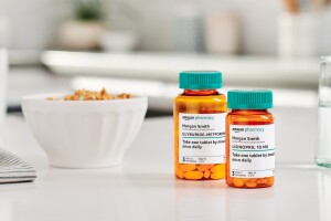 Two Amazon Pharmacy pill bottles sit on a white kitchen countertop in between a glass of water and a bowl of cereal.