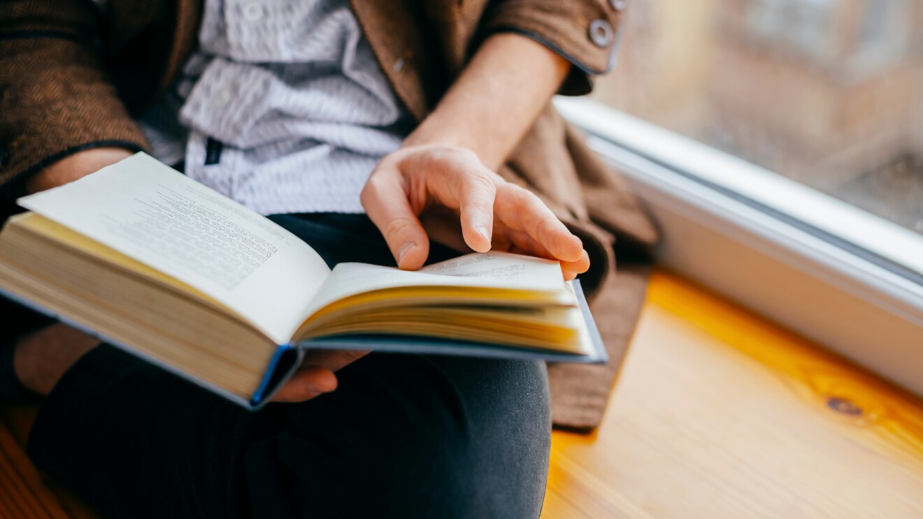 A photo of a person sitting on a window sill holding an open book in their lap.