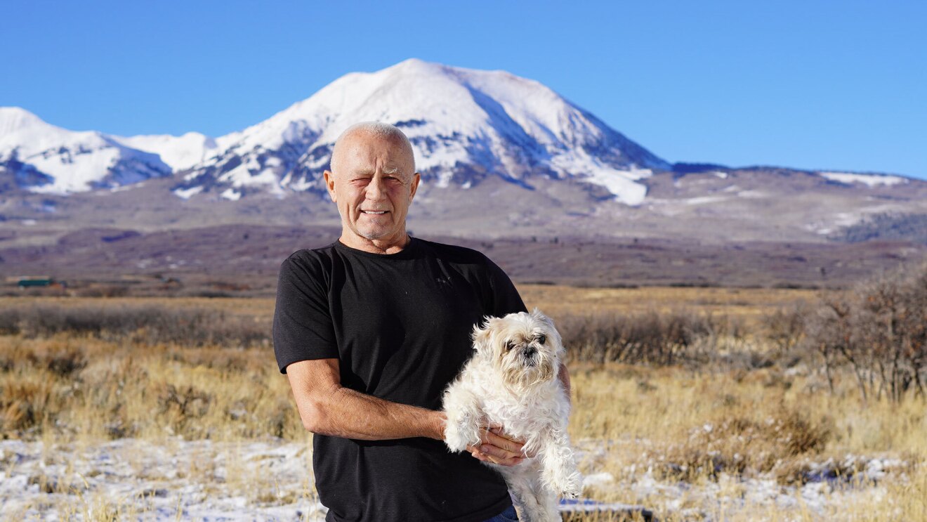 An image of a man smiling while holding a small white dog. There is a landscape behind him with a large, snow-capped mountain in the distance.