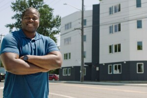 A photo of a man standing outside on a street crossing his arms. Behind him is an apartment building.