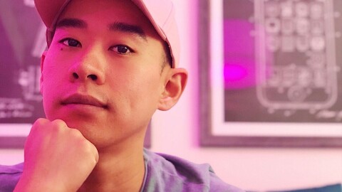 A headshot image of a man smiling softly for a photo under pink light. He has a white baseball hat on and has his chin rested on his fist.