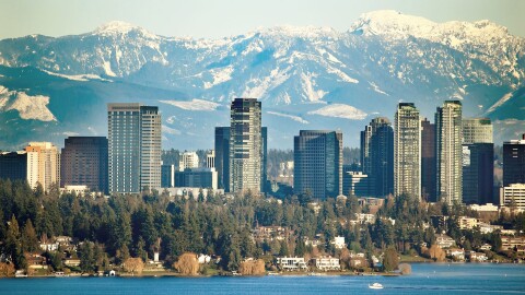 An image of the Bellevue, Washington skyline. There is a lake with trees on the shore in the foreground, tall buildings, and the Cascade mountain range in the background. 