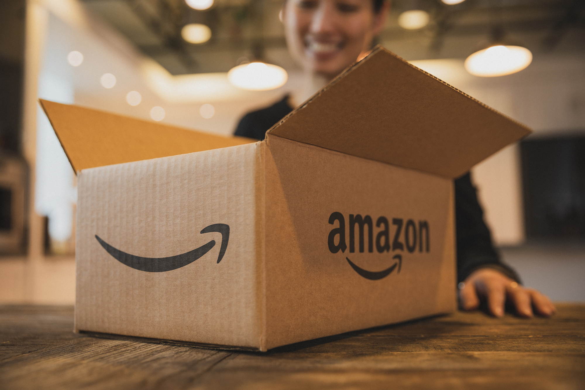 A smiling woman, slightly out of focus, stands behind an open shipping box emblazoned with the Amazon logo, which is resting on her kitchen table.