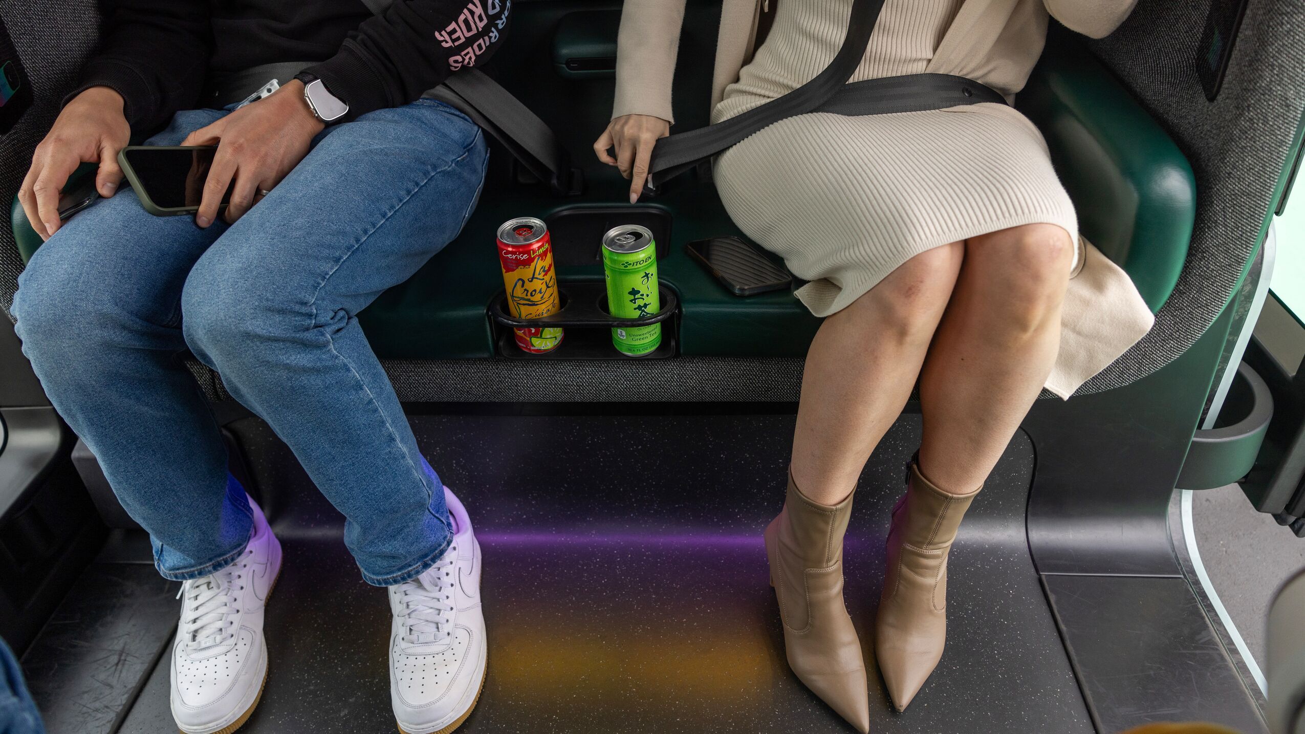 An image of two people's feet sitting in a Zoox robotaxi. There is a center console between them where they each have a drink in a can that is sitting in a cupholder.