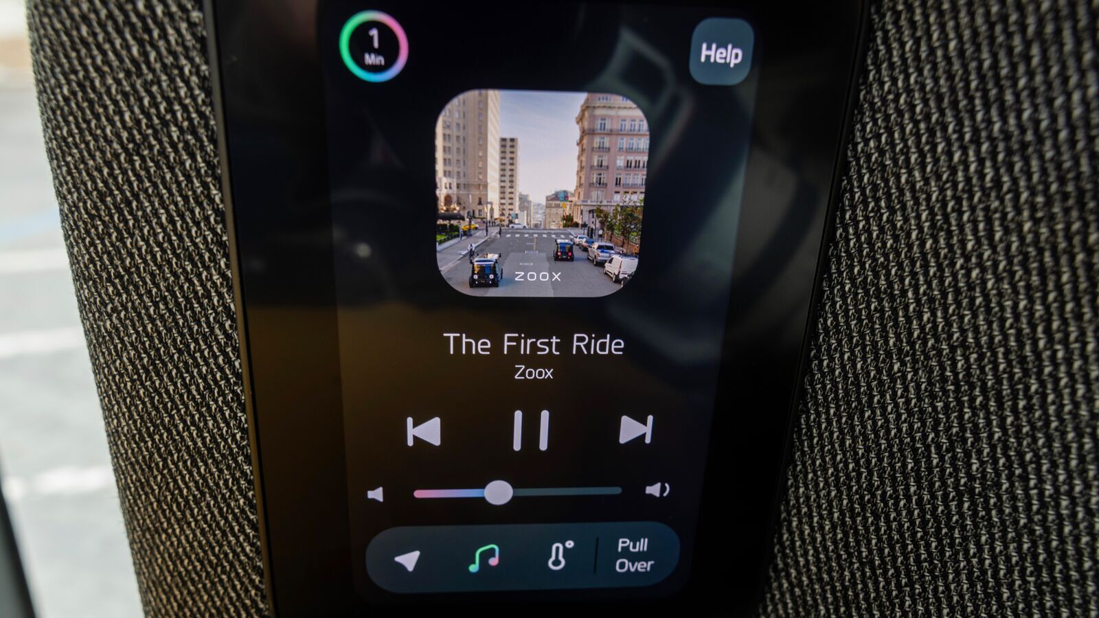 An image of a small screen inside the Zoox robotaxi. The screen shows an image of an album cover and the song title that says "The First Ride" with buttons to skip to the next song, pause, and turn up the volume.