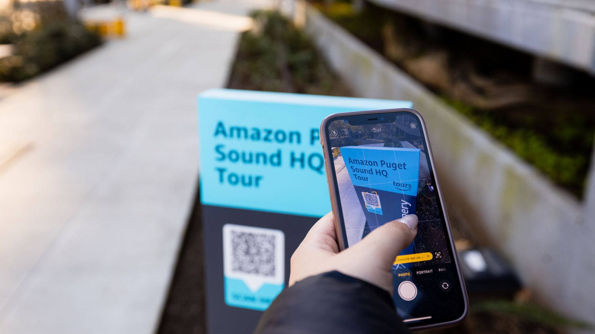 A photo of a person's hand holding a mobile device, taking a photo of the QR code displayed on a sign for the Amazon Puget Sound HQ Tour. 